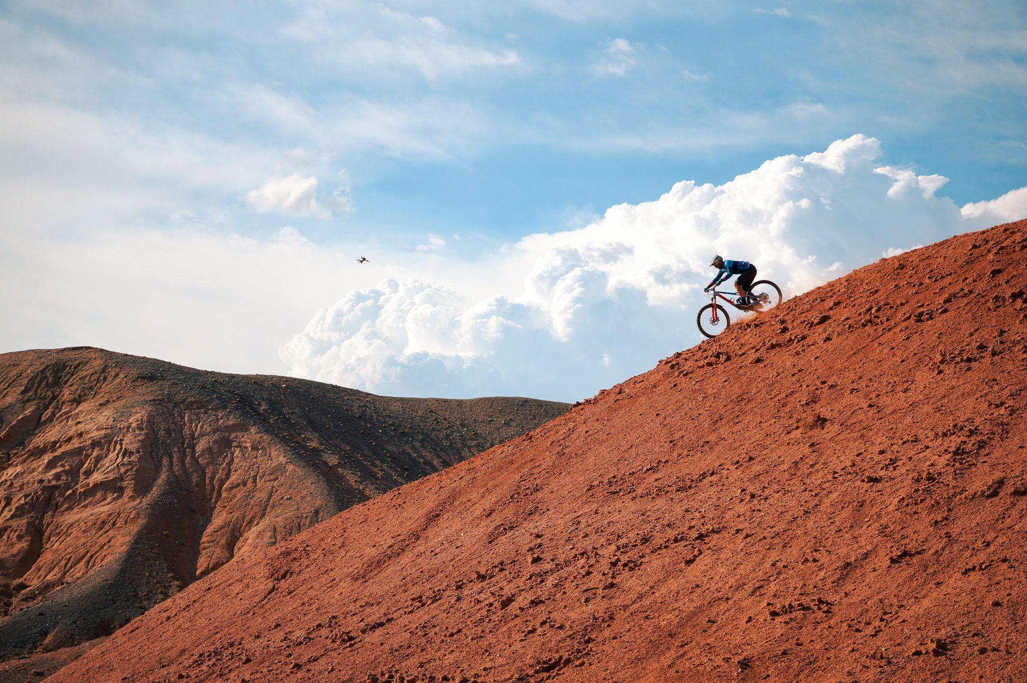 A mountain biker riding down a steep red ridge under tall white clouds